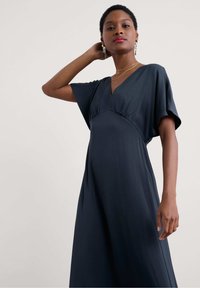 Sininen mekko, joka on valmistettu sileästä kankaasta; siinä on V-pääntie, lyhyet hihat sekä rento leikkaus ja ker gathered -yksityiskohta rinteen alla.