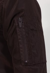 Giacca a vento marrone con tasca con zip sulla manica, texture liscia e vestibilità rilassata. Adatta per un abbigliamento casual.