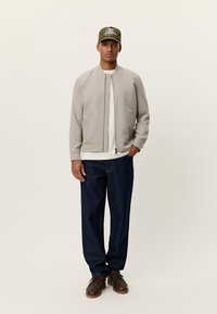 Φόρμα μπουφάν μπεζ ανοιχτού γκρι με φερμουάρ μπροστά, λευκό μπλουζάκι, σκούρο μπλε τζιν και καφέ παπούτσια. Το καπέλο έχει ένα γραφιστικό σχέδιο.