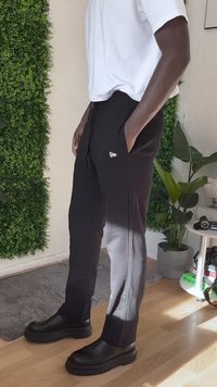 Person wearing black pants with white logo, white shirt, and black chunky boots, standing next to green plants and a small table with objects.