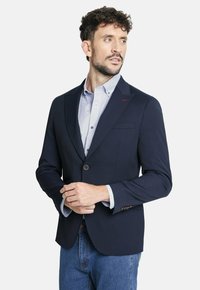 Blazer navy con un diseño estructurado, cierre de un solo botón, solapas con muesca y una camiseta de color azul claro que asoma de las mangas.
