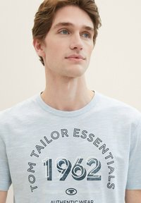 Svetlo modra majica s kratkimi rokavi in okroglim izrezom, izdelana iz mehkega blaga, s temno modrim in črnim grafičnim printom "TOM TAILOR ESSENTIALS 1962". Enostaven dizajn.