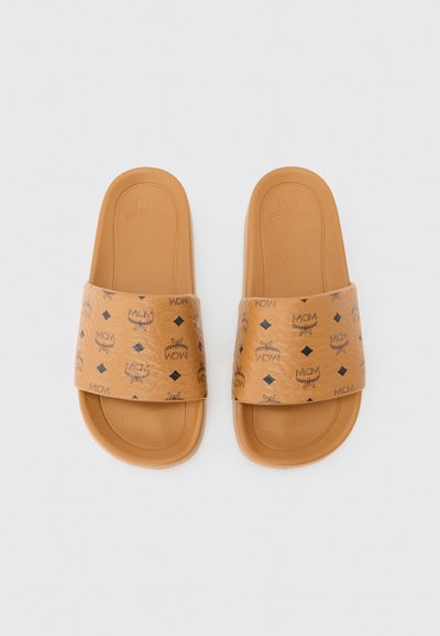 Paire de sandales slides couleur beige avec de larges brides ornées de motifs du logo MCM en noir, présentées sur un fond blanc uni.