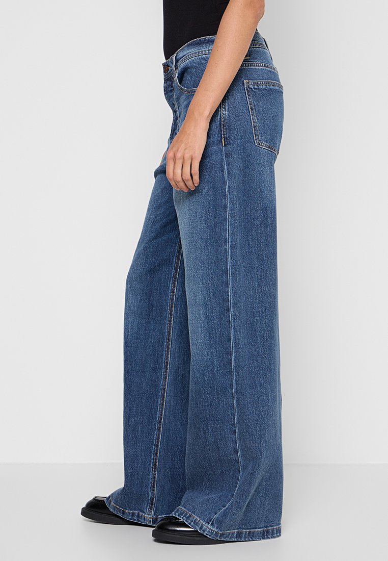 Frau, die hoch taillierte weit geschnittene blaue Jeans und schwarze Schuhe trägt, steht seitlich vor einem schlichten weißen Hintergrund.