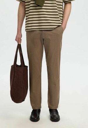 Homme portant un pantalon beige, une chemise à manches courtes rayée, des chaussures noires, tenant un sac cabas marron foncé dans une main, l'autre main dans la poche.