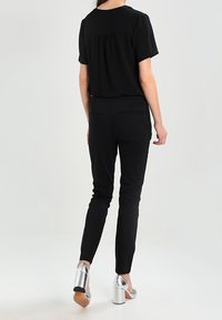Haut noir à manches courtes avec des plis dans le dos, associé à un pantalon noir ajusté et des chaussures à talons carrés en argent brillant.