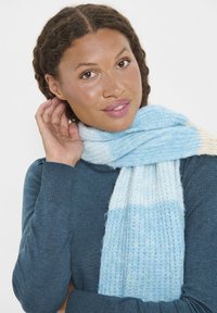 Sciarpa lavorata a maglia di color azzurro chiaro e crema, con una texture morbida, caratterizzata da un motivo a gradiente e bordi frangiati, appesa attorno al collo di una persona.
