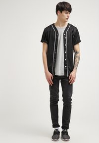 Chemise noire à manches courtes avec des bordures blanches, boutonnée sur le devant, portée par-dessus un t-shirt gris. Le modèle porte un jean noir déchiré et des baskets grises.