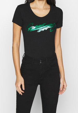 Woman wearing a black fitted t-shirt with green and white abstract text design, paired with high-waisted black jeans.