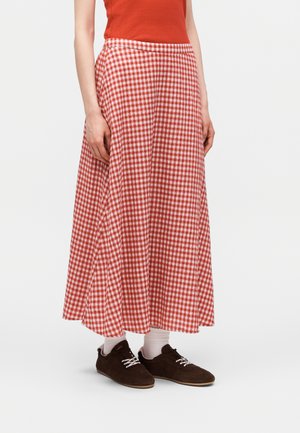 Žena v červené a bílé kostkované dlouhé sukni, červeném tílku bez rukávů, bílých ponožkách a tmavě hnědých šněrovacích botách na bílém pozadí.