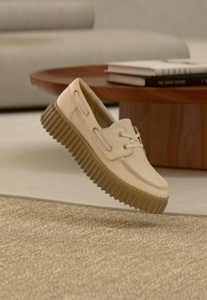 Beżowy skórzany mokasyn z grubą, ryflowaną gumową podeszwą unosi się nad teksturowanym beżowym dywanem, w tle drewniany stolik kawowy i sofa.