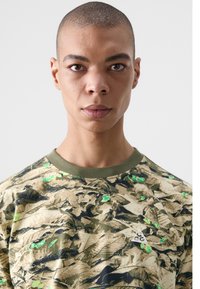 Camouflage-patterned sweatshirt in beige with green accents, featuring a crew neckline and a small white logo on the left side.