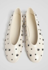 Cremefarvede læderballetsko prydet med rundede sølvfarvede nitte i forskellige størrelser, vist fra oven på en ensfarvet baggrund.