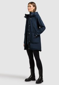 Femme portant un parka bleu marine à col montant, des leggings noirs et des bottines noires, debout de profil en trois quarts sur un fond uni.