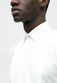 Fehér ing pliszírozott elejű, klasszikus gallérral és gombos rögzítéssel. Simább anyag és testre szabott szabás látható.