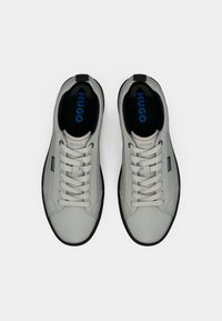 Αθλητικά παπούτσια σε ανοιχτό γκρι με λεία υφή, μαύρη σόλα, κορδόνια και μπλε λογότυπο "HUGO" στην επένδυση και τη γλώσσα.