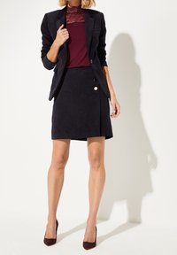 Blazer en velours côtelé bleu marine avec manches retroussées, associé à un haut en dentelle bordeaux et une jupe assortie bleu marine avec un détail de boutons sur le côté.