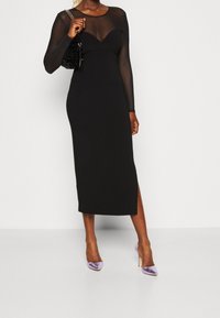 Czarna midi sukienka z przezroczystymi długimi rękawami, dopasowaną górą i bocznym rozcięciem. Połączona z fioletowymi satynowymi szpilkami i teksturowaną czarna torebką.