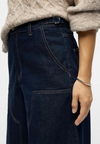 Beige gestrickter Pullover, in dunkelblaue Jeans mit großen Vordertaschen gesteckt, getragen mit einem silbernen Armband am rechten Handgelenk.