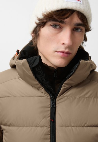 Tan puffer jacket with a high collar and black lining, featuring a zipper front, adjustable hood, and orange accent. Knitted white beanie.