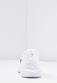 Bílé atletické boty s texturovaným vrškem, designem s křížovými popruhy, polstrovaným límcem a vroubkovou podešví. Logo na boku v černé barvě.