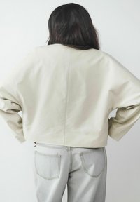 Skórzana jasnobeżowa kurtka o szerokich rękawach, wykonana z gładkiego materiału, w zestawieniu z jasnoszarymi dżinsami z tylnymi kieszeniami.