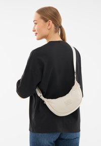 Borsa a spalla beige realizzata in tessuto morbido, forma curva, con tracolla regolabile, dotata di una piccola patch con logo. Il modello indossa una maglietta nera larga.