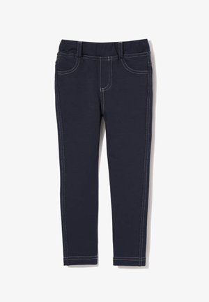 Uśmiechnięta dziewczyna z długimi brązowymi włosami, ubrana w białą bluzkę z długim rękawem, ciemne rurki i białe trampki, stojąca na jednej nodze.