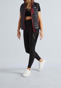 Schwarze, puffy Weste mit roten Akzenten über einem schwarzen Crop-Top und schwarzen Leggings, kombiniert mit weißen Sneakern. Glatter, enganliegender Stoff überall.