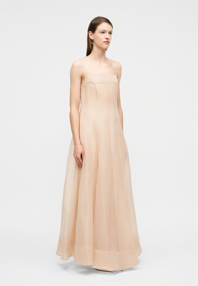 Γυναίκα που φοράει αμάνικο φόρεμα μέχρι το πάτωμα σε γυμνό χρ ώμα με λεπτές τιράντες, στέκεται μπροστά σε απλό λευκό φόντο.