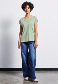Světle zelené tričko s krátkým rukávem a výstřihem ve tvaru lodi, doplněné texturovaným akcentem. Vzhled doplňují široké modré džíny a krémové boty.