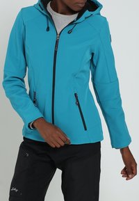 Personne portant une veste à capuche couleur sarcelle avec une fermeture éclair avant et des poches zippées, associée à un pantalon noir, debout devant un fond uni.