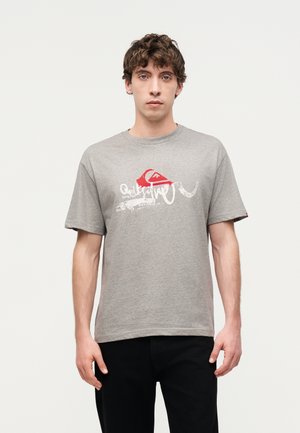 Młody mężczyzna z krótkimi kręconymi włosami nosi szary T-shirt z nadrukiem i czarne spodnie, stoi na tle jednolitego jasnego tła.