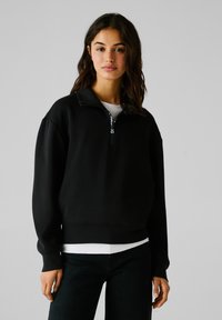 Sudadera negra de pulóver con cuello alto con cremallera, que presenta puños acanalados y un corte relajado. El material parece suave y liso al tacto.