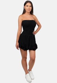 Zwarte strapless jurk met een gesmokte boezem en gerimpelde rok. Model draagt witte sneakers. Glad stoffen textuur, aansluitend ontwerp.