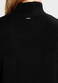 Schwarzer Strickpullover mit hohem Kragen und weicher Textur. Enthält ein kleines metallisches Markenschild auf der Rückseite.
