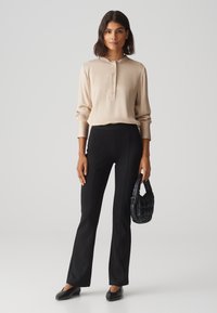 Blouse beige à manches longues avec un col mandarin et patte de boutonnage, associée à un pantalon noir évasé et un sac à main noir texturé.