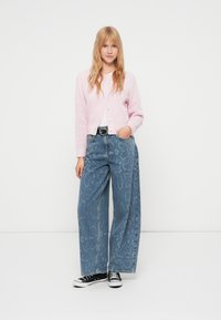 Ανοιχτό ροζ πλεκτή ζακέτα με κουμπιά, συνδυασμένη με φαρδιά μπλε τζιν παντελόνια με διακριτικό σχέδιο και μαύρα αθλητικά παπούτσια.