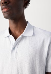 Biely polo tričko s textúrovaným dizajnom, dvoma svetlomodrými gombíkmi a jemne vyšitým logom na hrudi. Mäkký bavlnený materiál.