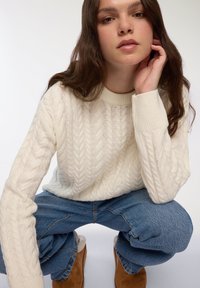 Kremowy sweter w warkocz z okrągłym dekoltem i ściągaczami, w połączeniu z niebieskimi spodniami z wysokim stanem i beżowymi botkami do kostki.