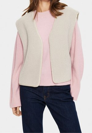 Vrouw draagt een lichtbeige mouwloos vest met open voorkant over een roze geribbelde trui en donkerblauwe jeans tegen een witte achtergrond.
