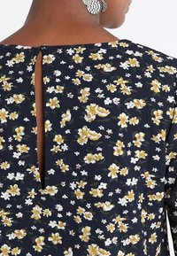 Granatowa bluzka w kwiaty z małymi białymi i żółtymi wzorami, z detalem w formie dziurki z tyłu oraz teksturowanym materiałem.