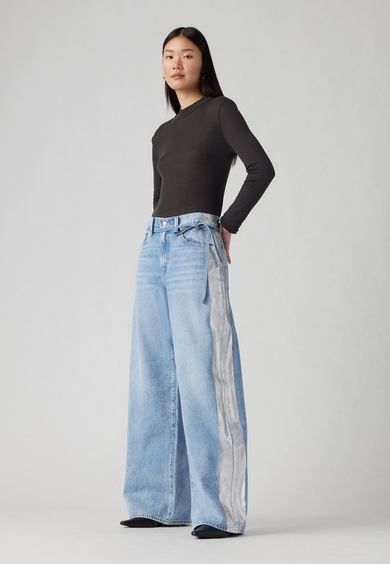 Jeans a gamba larga blu chiaro con pannello laterale argentato, vita alta e dettagli con cordino, abbinati a un top nero a maniche lunghe.