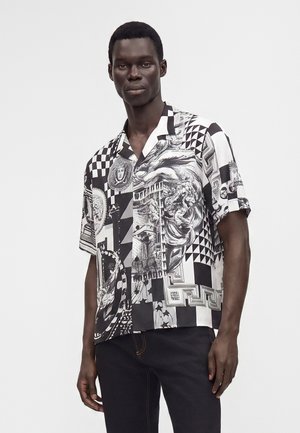 Man draagt een zwart-wit kortemouwen blouse met knopen met gemengde geometrische en klassieke kunstpatronen, gecombineerd met donkere broek.