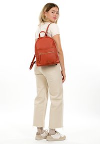 Jeune femme portant un pantalon beige et des baskets blanches, légèrement tournée avec un sac à dos en cuir couleur rouille sur le dos.