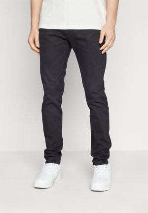 Unterer Teil einer Person, die eng anliegende schwarze Jeans, weiße Turnschuhe und ein weißes Hemd vor einem schlichten hellen Hintergrund trägt.