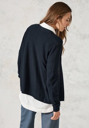 Marineblauwe gebreide cardigan met een gestructureerd ontwerp, met een relaxte pasvorm en geribde manchetten, gedragen over een witte button-up shirt.