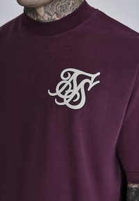 Camiseta color ciruela con un cuello alto y mangas cortas, que presenta un gran logotipo estilizado en blanco en el pecho y una textura suave y lisa.