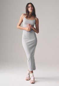 Szara, dopasowana, długa sukienka na cienkich ramiączkach z bocznym rozcięciem. Połączona z różowymi tenisówkami i białymi prążkowanymi skarpetkami do kostek. Gładkie tło.