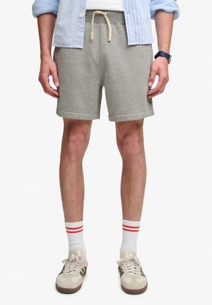 Hombre con pantalones cortos gris claro con cordón, calcetines blancos con rayas rojas, zapatillas beige, reloj azul y camisa azul a rayas con mangas remangadas.